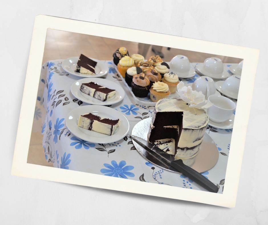 Table set for morning tea with sliced chocolate layer cake, assorted cupcakes, and white teacups on a floral tablecloth at Kirkley Gardens retirement village.