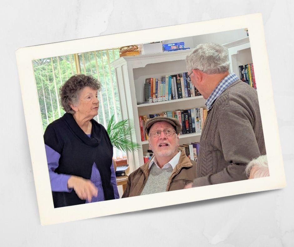 Three older adults chatting in the community centre at Kirkley Gardens retirement village in Lithgow, surrounded by bookshelves and natural light.