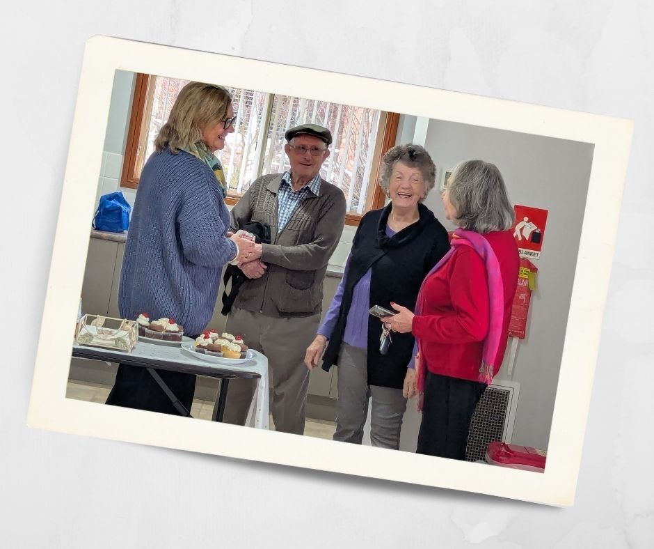 Four smiling Kirkley Gardens residents chatting and laughing together indoors at a community morning tea in Lithgow retirement village, with cupcakes on a nearby table.